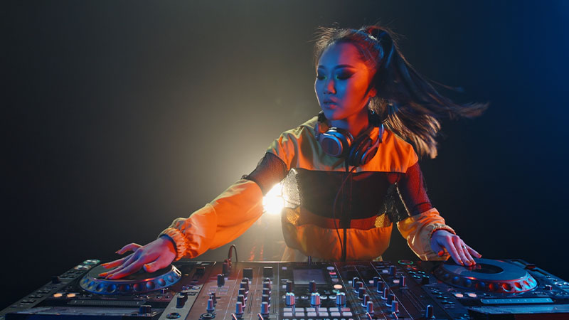 DJ performing to a crowd at an outdoor event, surrounded by trees and snow. People are watching and some are taking photos.