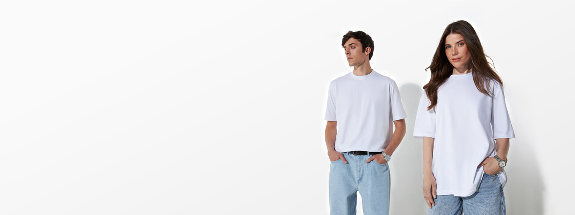 A man and a woman stand wearing oversized white t-shirts and blue jeans against a plain white background.