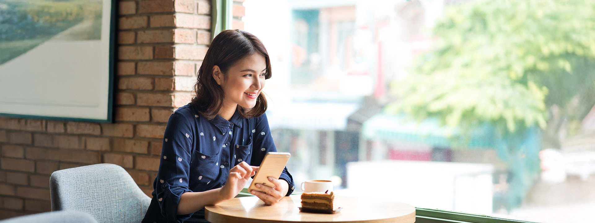 Woman sitting at a café table by a window, holding a smartphone, with a cup of coffee and a slice of cake in front of her.