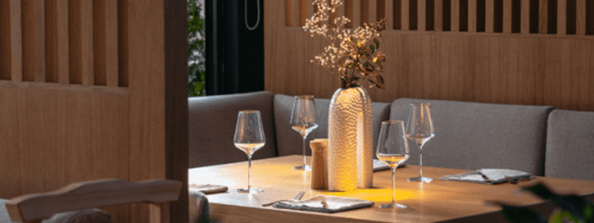 A wooden dining table set for four with wine glasses, cloth napkins, and a decorative vase with dried flowers in a modern restaurant setting.