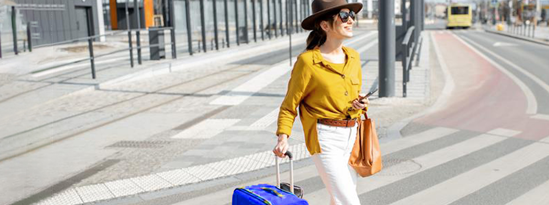 Woman in a yellow blouse and hat walks on a street, pulling a blue suitcase, holding a phone, with tram tracks and a tram in the background.