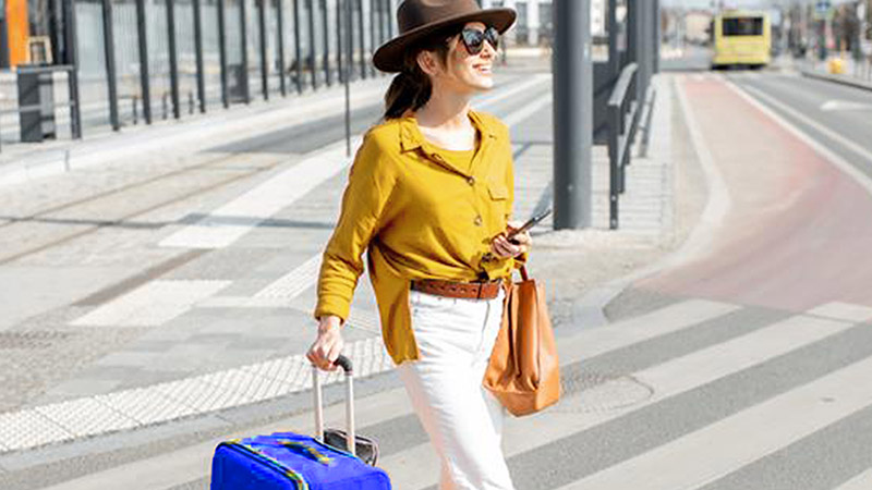 Woman in a yellow blouse and hat walks on a street, pulling a blue suitcase, holding a phone, with tram tracks and a tram in the background.