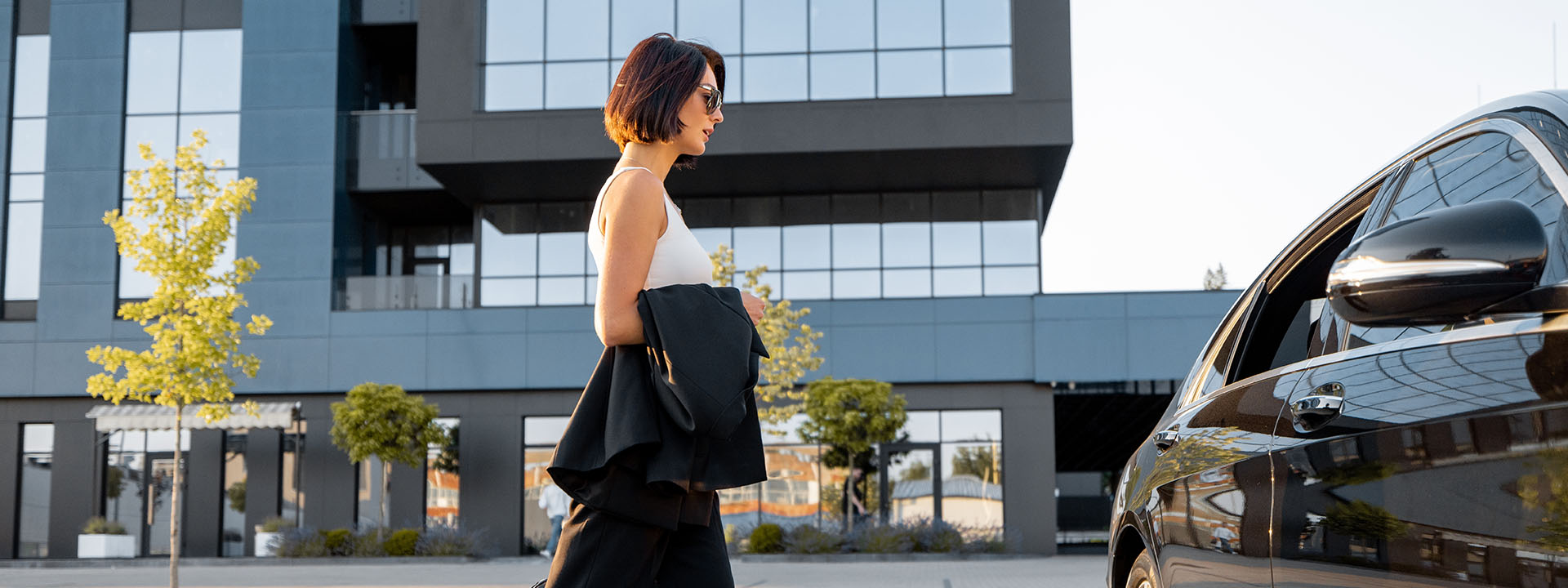 A woman in sunglasses walks toward a black car in front of a modern glass building on a sunny day.