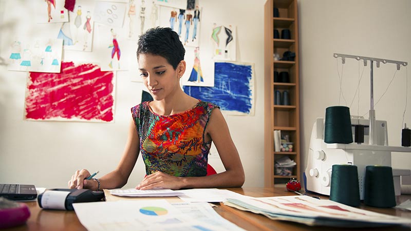 Small business owner sitting at desk using calculator, sketches hanging on wall behind her.