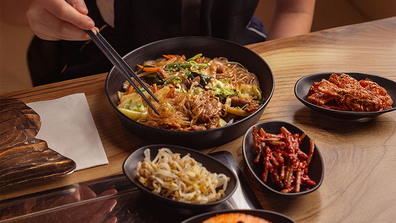 A person uses chopsticks to eat from a bowl of noodles with vegetables, surrounded by side dishes of kimchi, bean sprouts, and spicy strips on a wooden table.