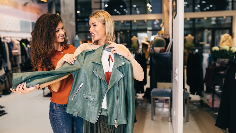 Two women in a store holding a green leather jacket, smiling and discussing the item. Clothing and accessories are displayed in the background.