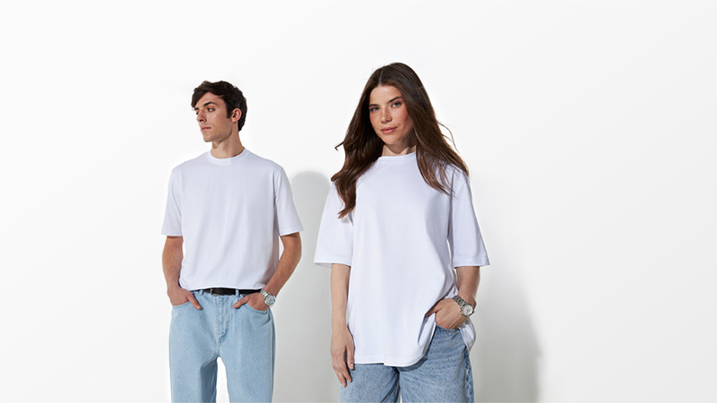 A man and a woman stand wearing oversized white t-shirts and blue jeans against a plain white background.