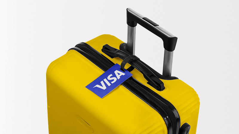 A yellow suitcase with a Visa tag attached to the handle against a plain background.