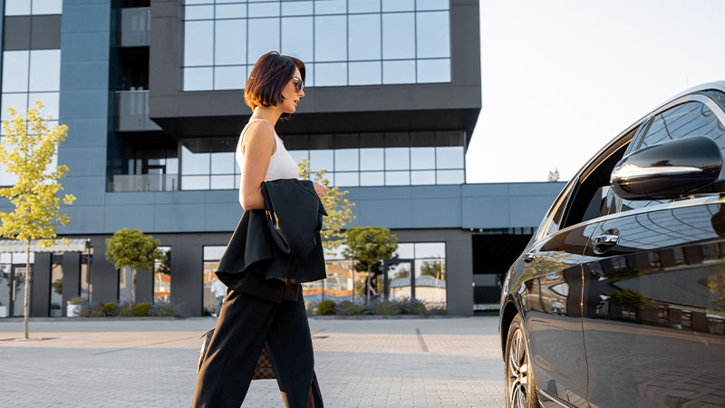 A woman in sunglasses walks toward a black car in front of a modern glass building on a sunny day.
